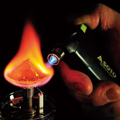 A hand holds the black Soto Outdoors Pocket Torch near a small stove with a bright flame.