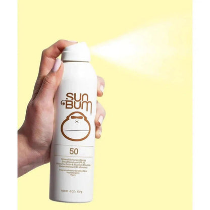 A hand holds a white Sun Bum Mineral SPF 50 Sunscreen Spray can spraying against a yellow background.