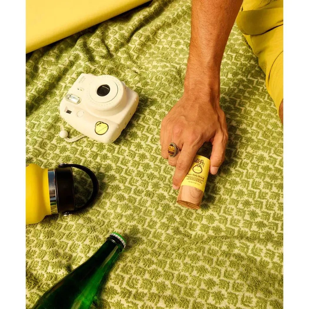 A hand holds the Sun Bum OG SPF 50 Face + Body Stick on a green patterned blanket with a white camera nearby.