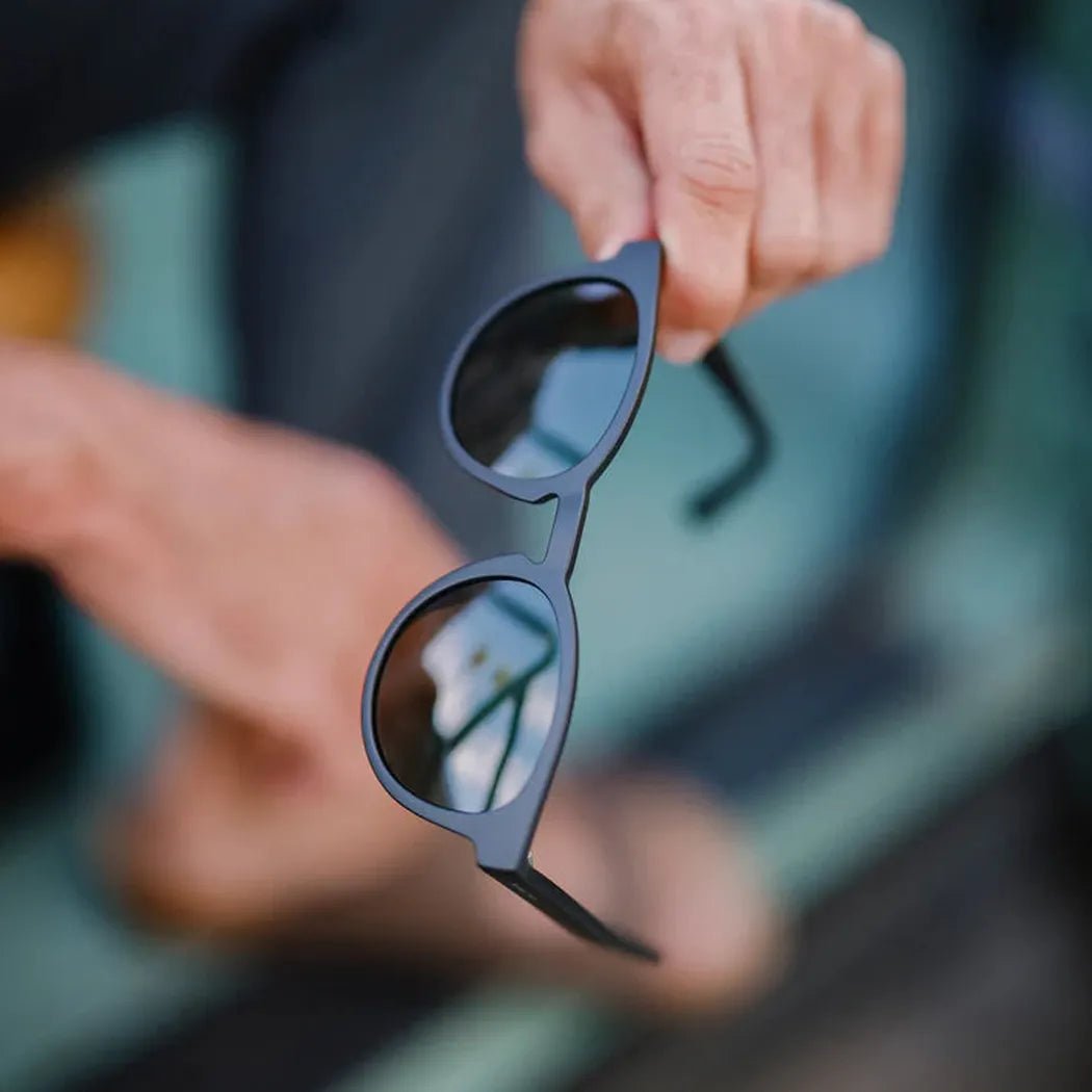 A hand holds the Suncloud Barnes sunglasses with matte black frames, showing the reflective lenses and thin arms against a blurred outdoor background.