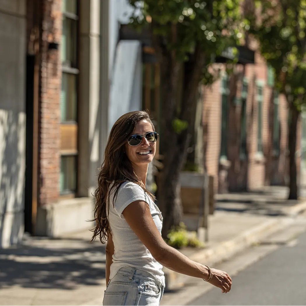 A woman wearing Gold Suncloud Patrol sunglasses smiles while walking on a sunny city sidewalk.