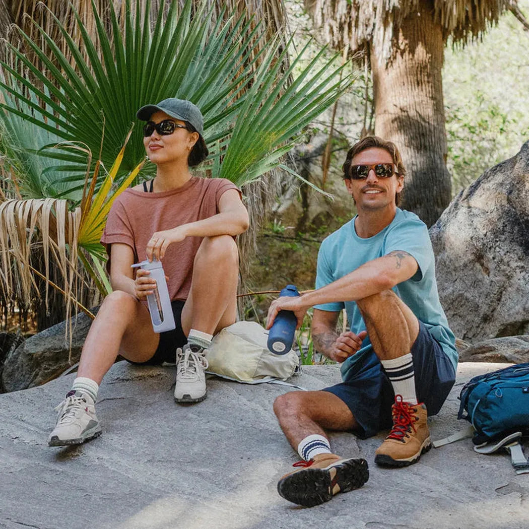 A woman and man relax outdoors wearing Suncloud Sculpin Matte Black sunglasses, with palm trees and rocks in the background.
