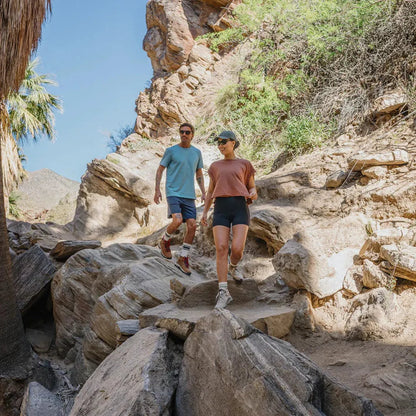 A man and woman hike on rocky terrain wearing Suncloud Sculpin Matte Black sunglasses, with palm trees and a clear blue sky in the background.