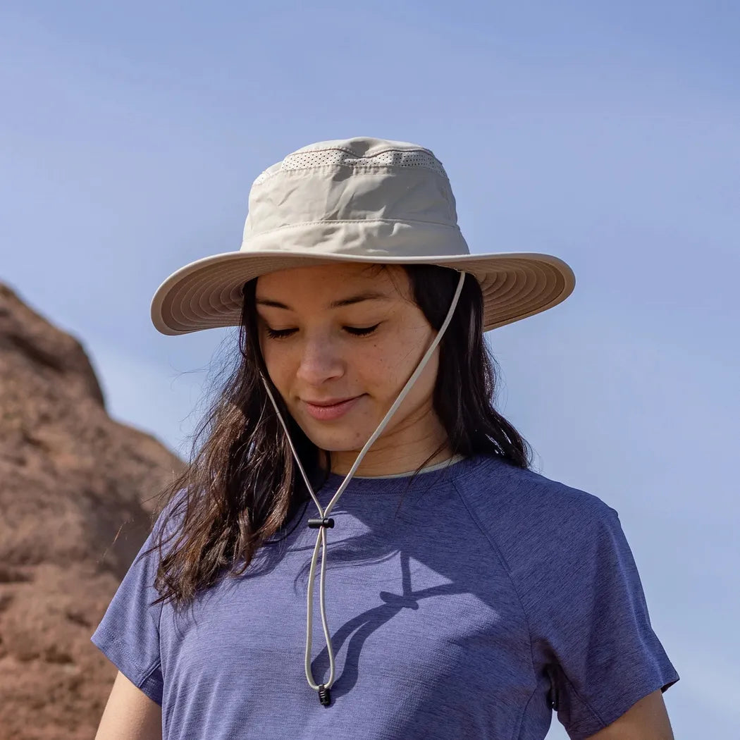 A woman wears the Sunday Afternoons Cruiser Hat in Cream/Sand, with a wide brim and chin strap, against a clear blue sky and rocky background.