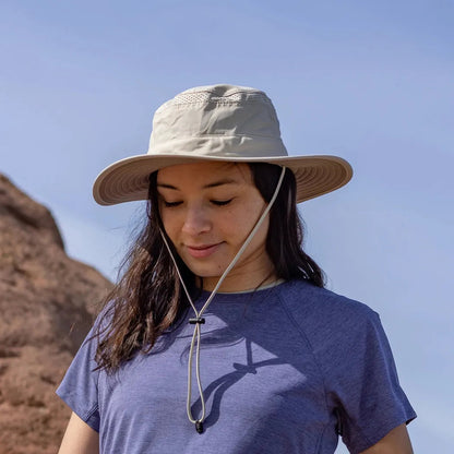 A woman wears the Sunday Afternoons Cruiser Hat in Cream/Sand, with a wide brim and chin strap, against a clear blue sky and rocky background.