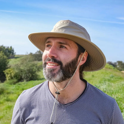 A man wears the Sunday Afternoons Cruiser Hat in Cream/Sand while standing in a grassy field under a blue sky.