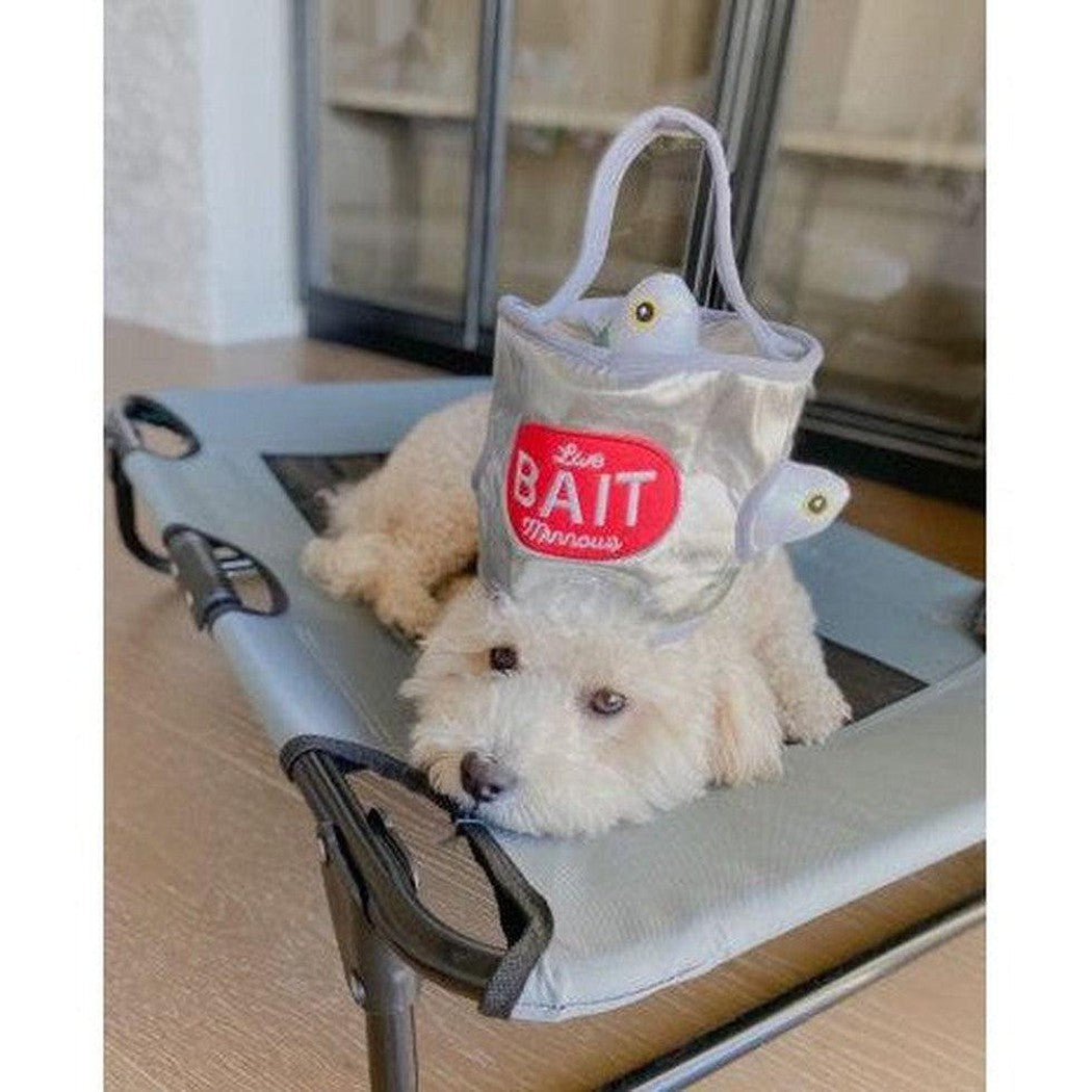 A white dog wears the Tall Tails Bait Bucket Squeaky Hide & Seek Toy on a gray pet bed with a glass background.