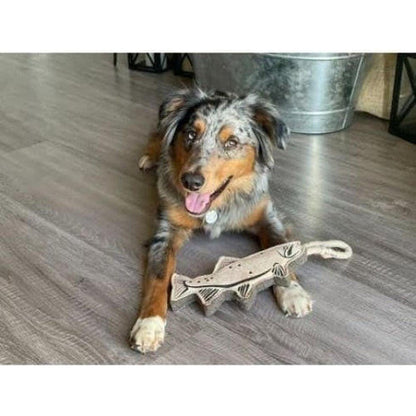 A dog lies on a wood floor with the Tall Tails Natural Leather Trout Tug Toy in front of it, near a metal bucket.