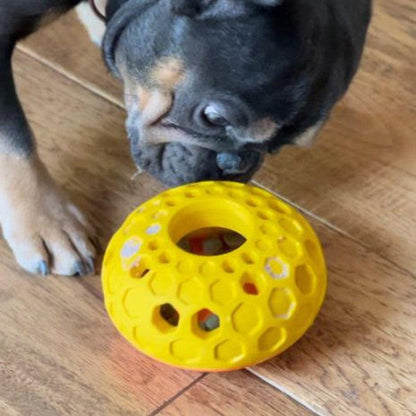 A black and tan dog sniffs a yellow Tall Tails Natural Rubber Bee Hive Toy on a wooden floor.
