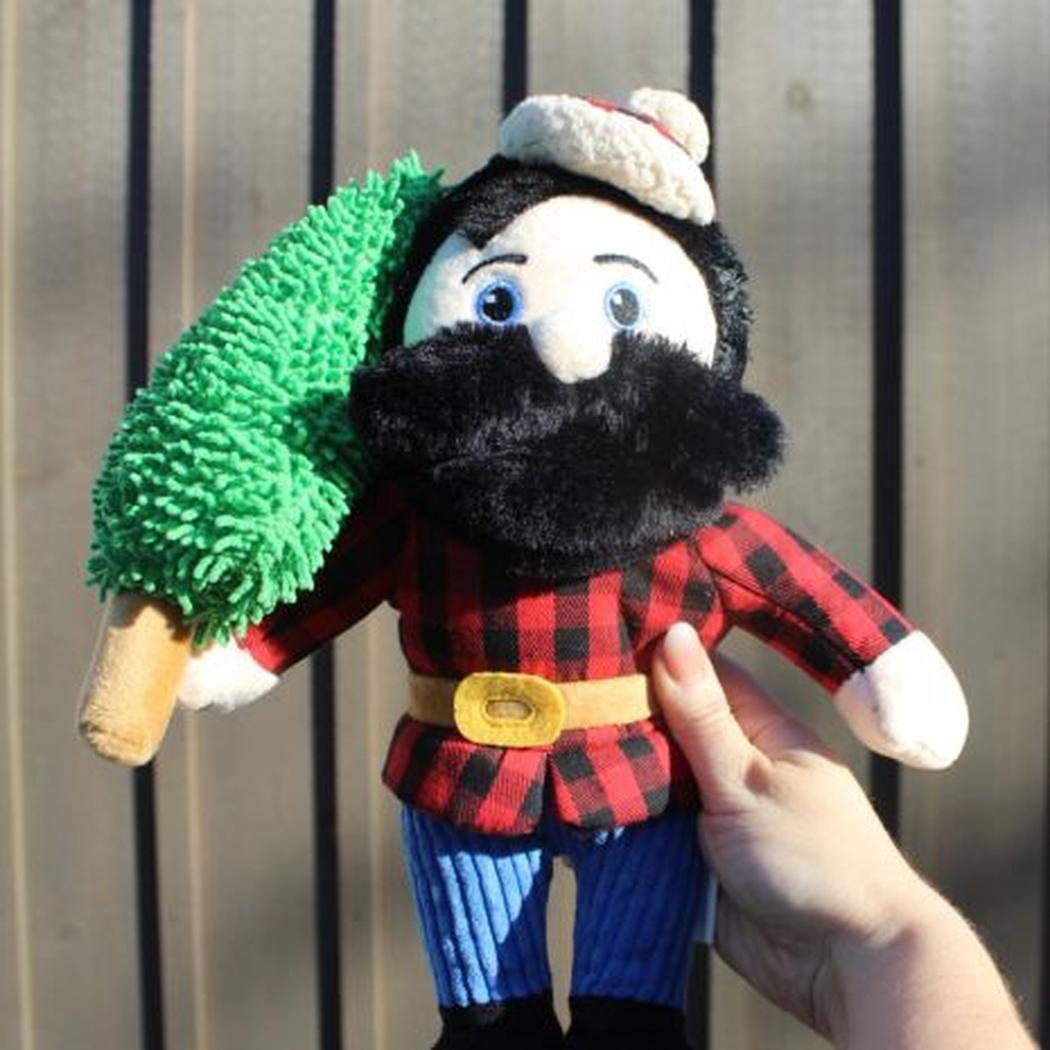 A hand holds the Tall Tails Paul Bunyan Rope Body Dog Toy, showing its red and black plaid shirt, blue pants, and green tree accessory against a wooden background.