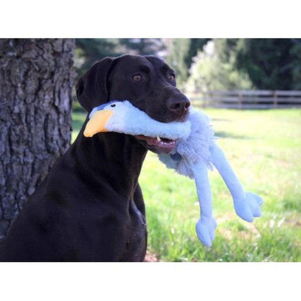 A black dog holds the Tall Tails Plush Rope Body Heron Squeaker Toy in its mouth while sitting by a tree in a grassy field.