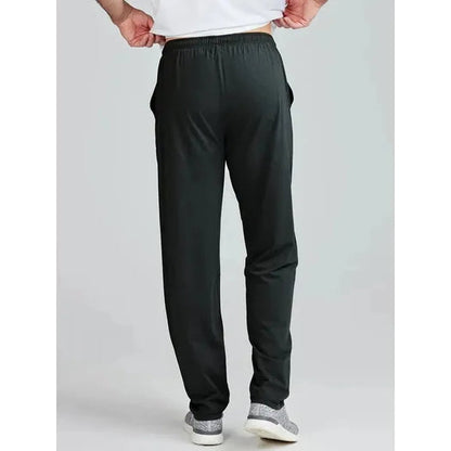 Back view of the Tasc Men's Carrollton Classic Pant in Black on a model wearing gray sneakers against a light gray background.