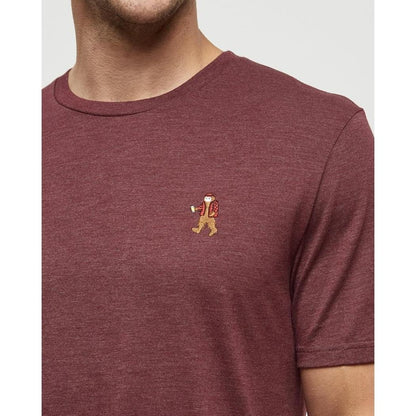 A close-up of a man wearing the Tentree Men's Sasquatch T-Shirt in Deep Burgundy Heather Lumberjack, showing the small embroidered Sasquatch logo on the chest against a plain background.