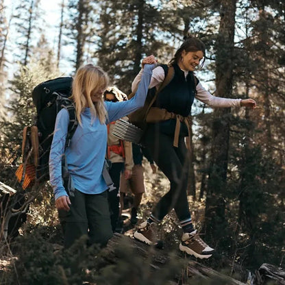Two women hike in a forest, one wearing Teva Women's Grandview Max GTX boots in Honey Brown/ Birch, helping the other step on a log.