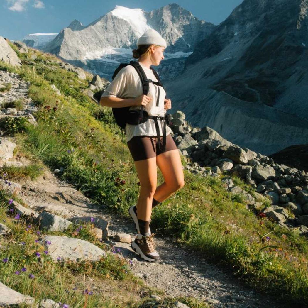 A woman hikes on a mountain trail wearing the Honey Brown/ Birch Teva Women's Grandview Max GTX boots, with snow-capped peaks in the background.