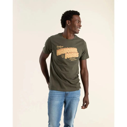 A model wears The Landmark Project Greetings From the National Parks Unisex Short Sleeve Tee in Dark Oak against a plain background.