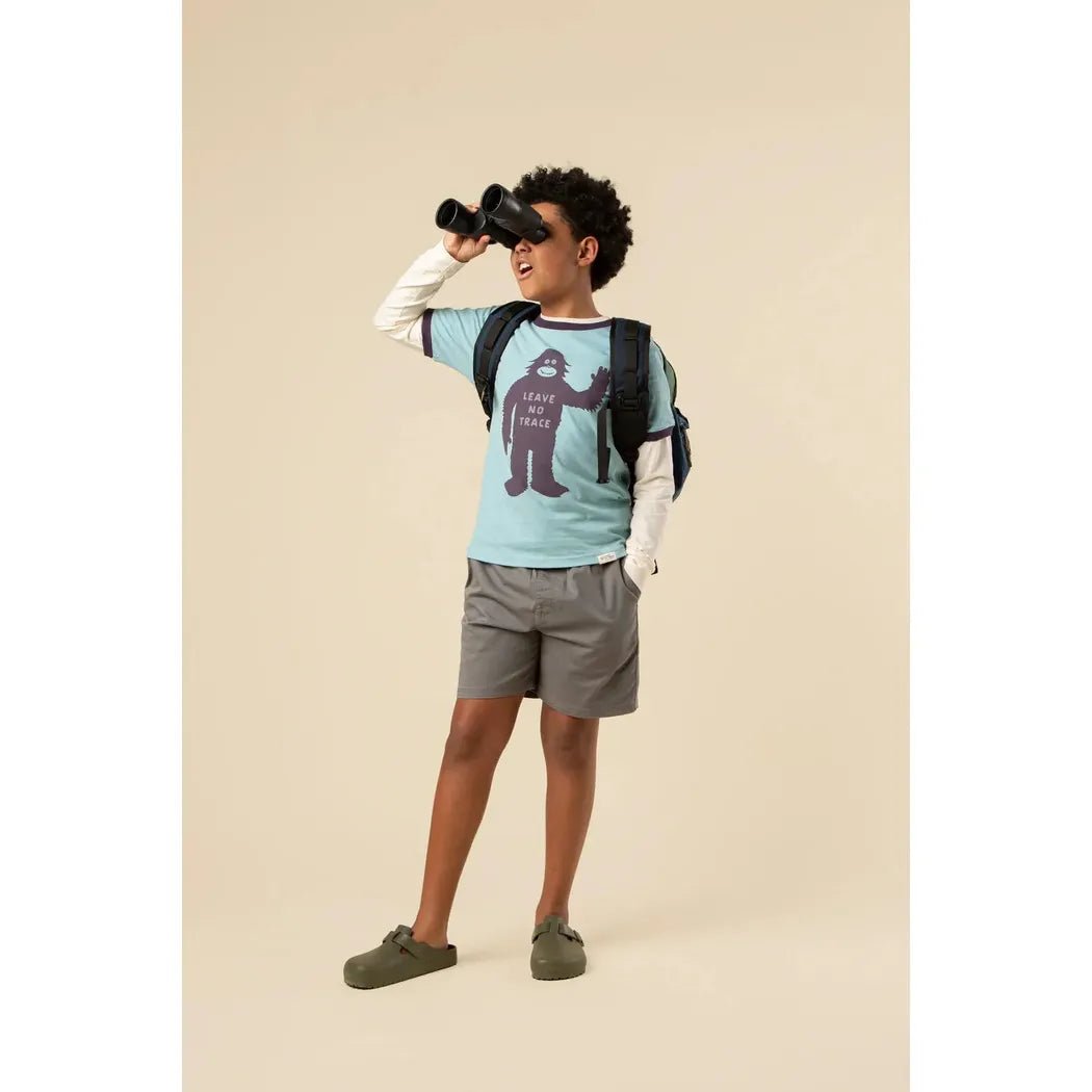 A young model wearing The Landmark Project Leave No Trace Youth Short Sleeve Ringer Tee in Glacier Blue/Dusk looks through binoculars against a neutral background.