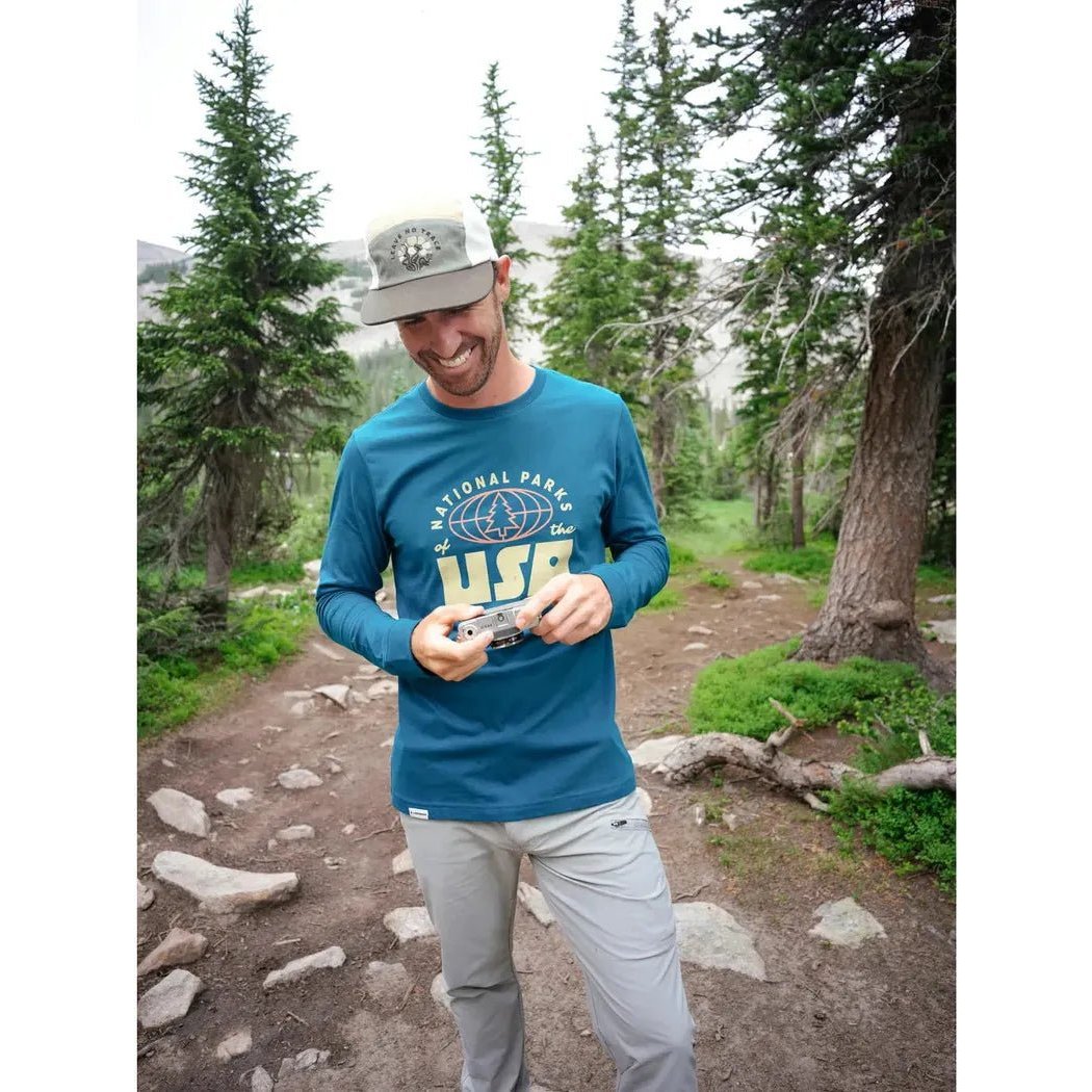 A man in a Vintage Royal The Landmark Project National Parks of the USA Unisex Long Sleeve Tee stands on a forest trail holding a camera.