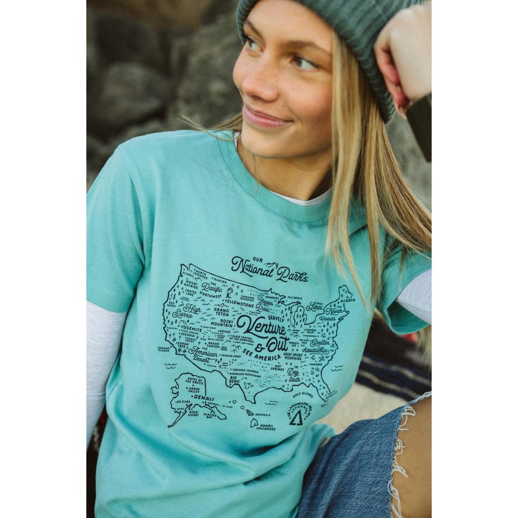 A model wears The Landmark Project NPS Map Youth Tee in Glacier Blue with a map graphic, against a rocky background.