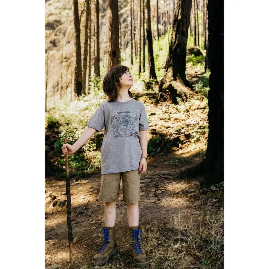 A youth wearing The Landmark Project NPS Map Youth Tee in Glacier Blue stands in a forest with a hiking pole.