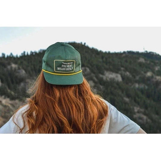 A woman with long red hair wears The Landmark Project Only You Hat, looking out at a forested mountain landscape.
