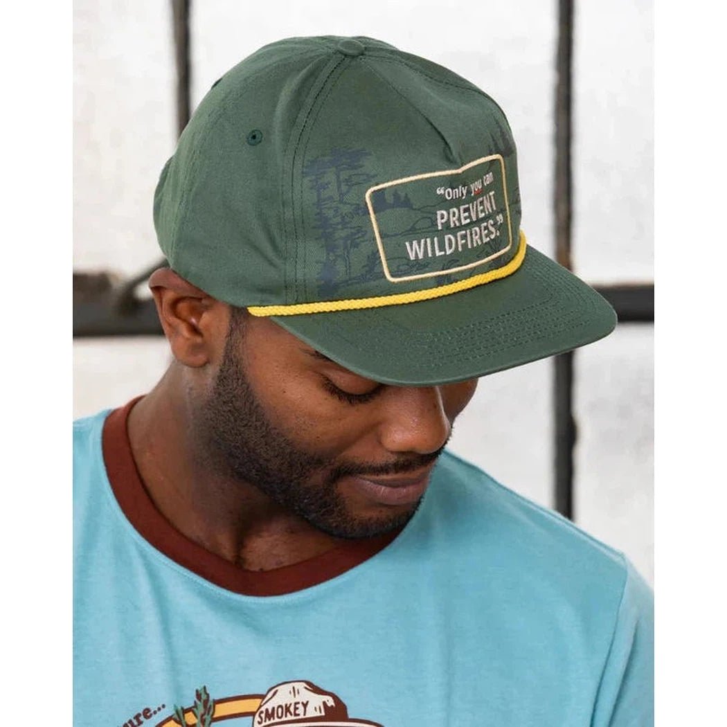 A man wears The Landmark Project Only You Hat in green with a yellow brim and a patch that says "PREVENT WILDFIRES" against a window background.