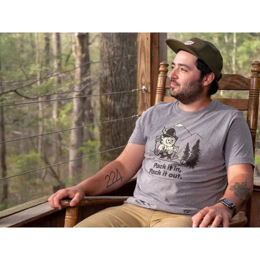 A man in a Smoke Grey The Landmark Project Pack It in, Pack It Out t - shirt sits on a wooden chair on a porch with trees in the background.