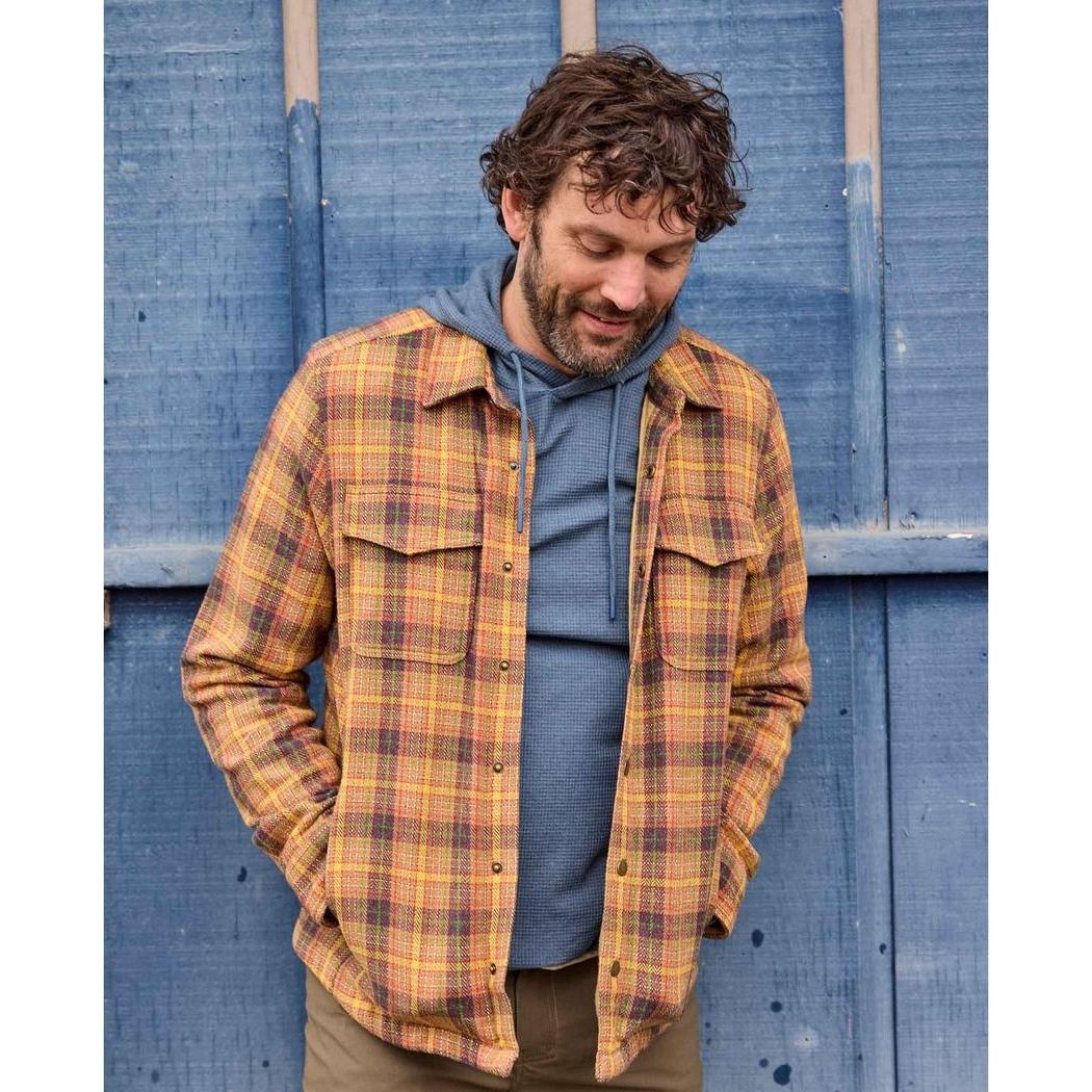 A man wearing the Toad & Co Men's Framer II Long Sleeve Hoodie in Pacific Blue stands with his hands in his pockets against a blue wooden background.
