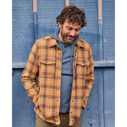 A man wearing the Toad & Co Men's Framer II Long Sleeve Hoodie in Pacific Blue stands with his hands in his pockets against a blue wooden background.