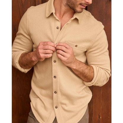 A man wearing the Toad & Co Men's Shadowbrook Long Sleeve Shirt in Camel Twill buttons it up against a wooden background.