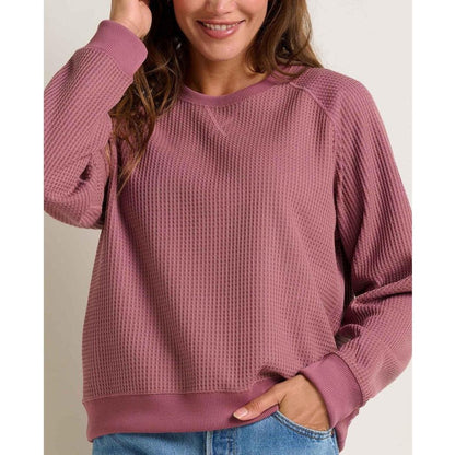 A woman wears the Toad & Co Women's BitterRoot Long Sleeve Pullover in Foxglove, showing the front and a hand in a pocket against a plain background.