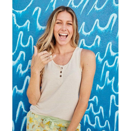 A woman wearing the Toad & Co Women's Piru Henley Tank in Salt poses against a blue wave-patterned background.