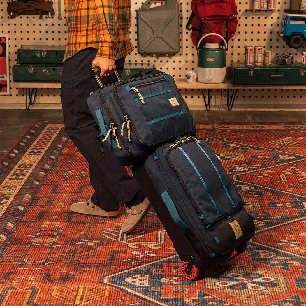 A person pulls the Topo Designs Global Travel Bag 30L in Navy / Sahara on a patterned rug with a pegboard background.