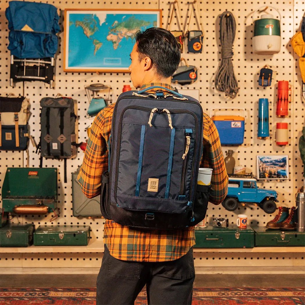 A man in a plaid shirt wears the navy Topo Designs Global Travel Bag 30L in front of a pegboard wall with travel gear.
