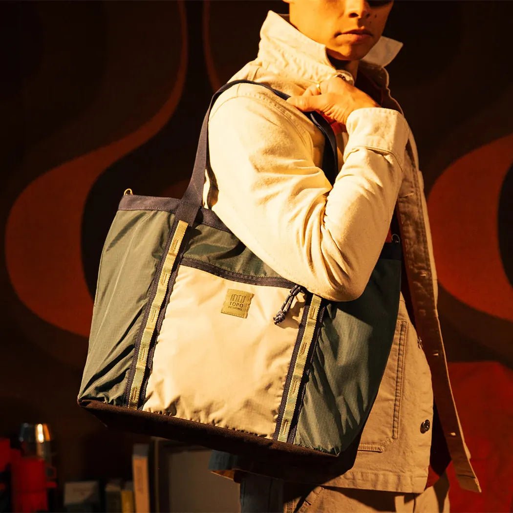 A model wears the Topo Designs Mountain Utility Tote in Forest / Bone White, showing its large size and shoulder strap against a warm, patterned background.