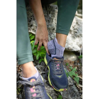 A woman in green leggings adjusts her black Tough Cuties Eve The Original Ladies Light Weight Hiker No Show Tab socks while wearing charcoal/gray hiking shoes on rocky ground.