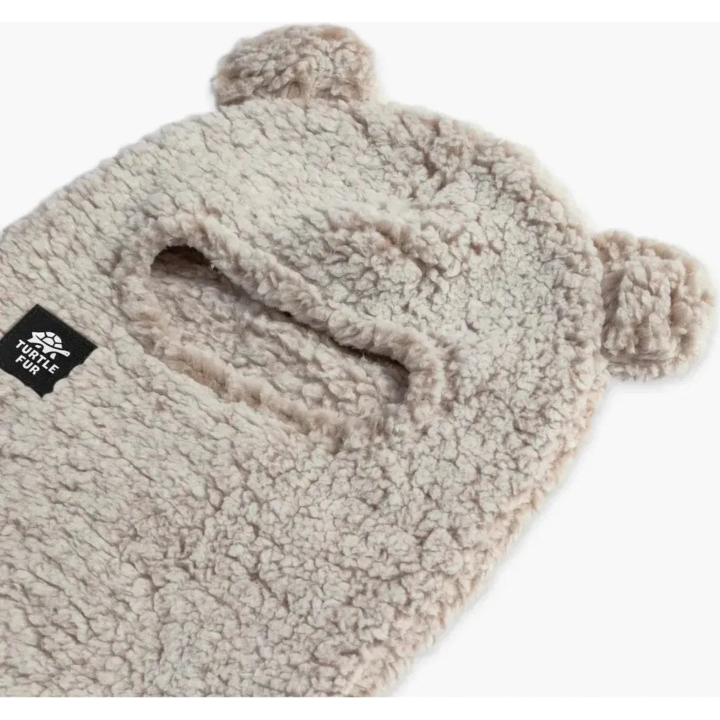 A natural Turtle Fur Kids Comfort Lush Bear Balaclava with a pom pom and ear flaps, shown from a top-down angle on a white background.