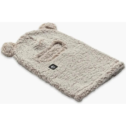 A natural Turtle Fur Kids Comfort Lush Bear Balaclava with two pom-pom ears and a small black tag, laid flat against a white background.