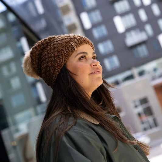 A woman wears the Turtle Fur Women's Snowfall hat in Camel, looking up at a city building.