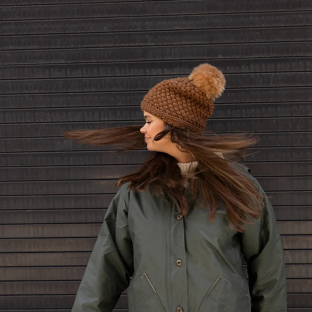 A woman wears the Turtle Fur Women's Snowfall hat in Camel, with her hair blowing in the wind against a dark background.