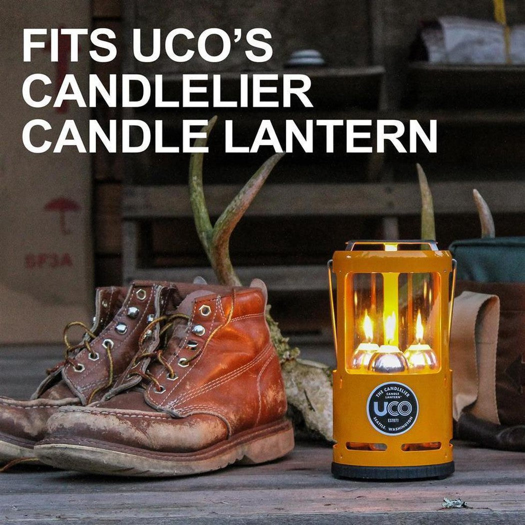 A yellow UCO candle lantern with lit candles sits next to brown work boots on a wooden surface.