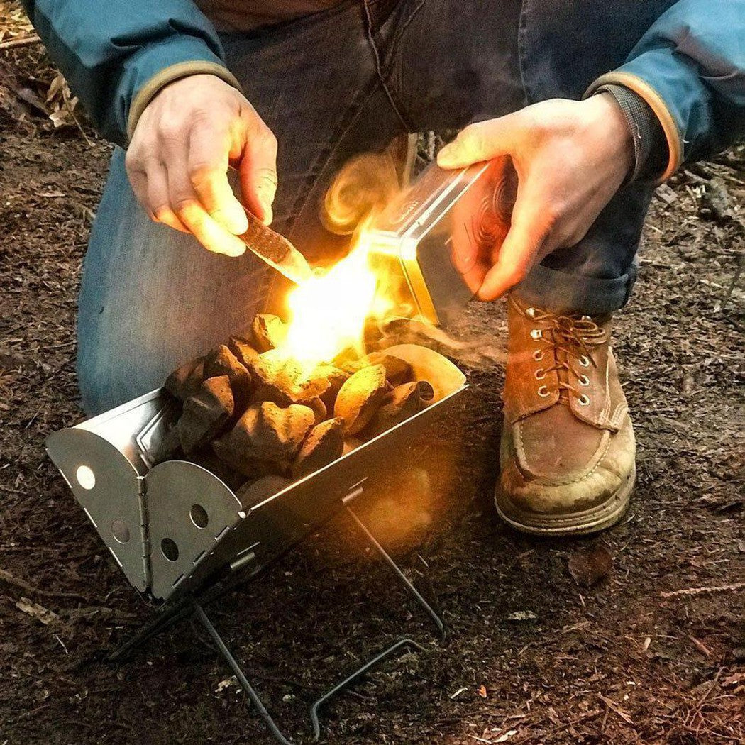 A person uses the UCO Behemoth Sweetfire Match Kit to light a small fire in a metal fire pit on the ground.