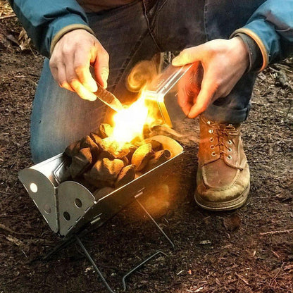 A person uses the UCO Behemoth Sweetfire Match Kit to light a small fire in a metal fire pit on the ground.