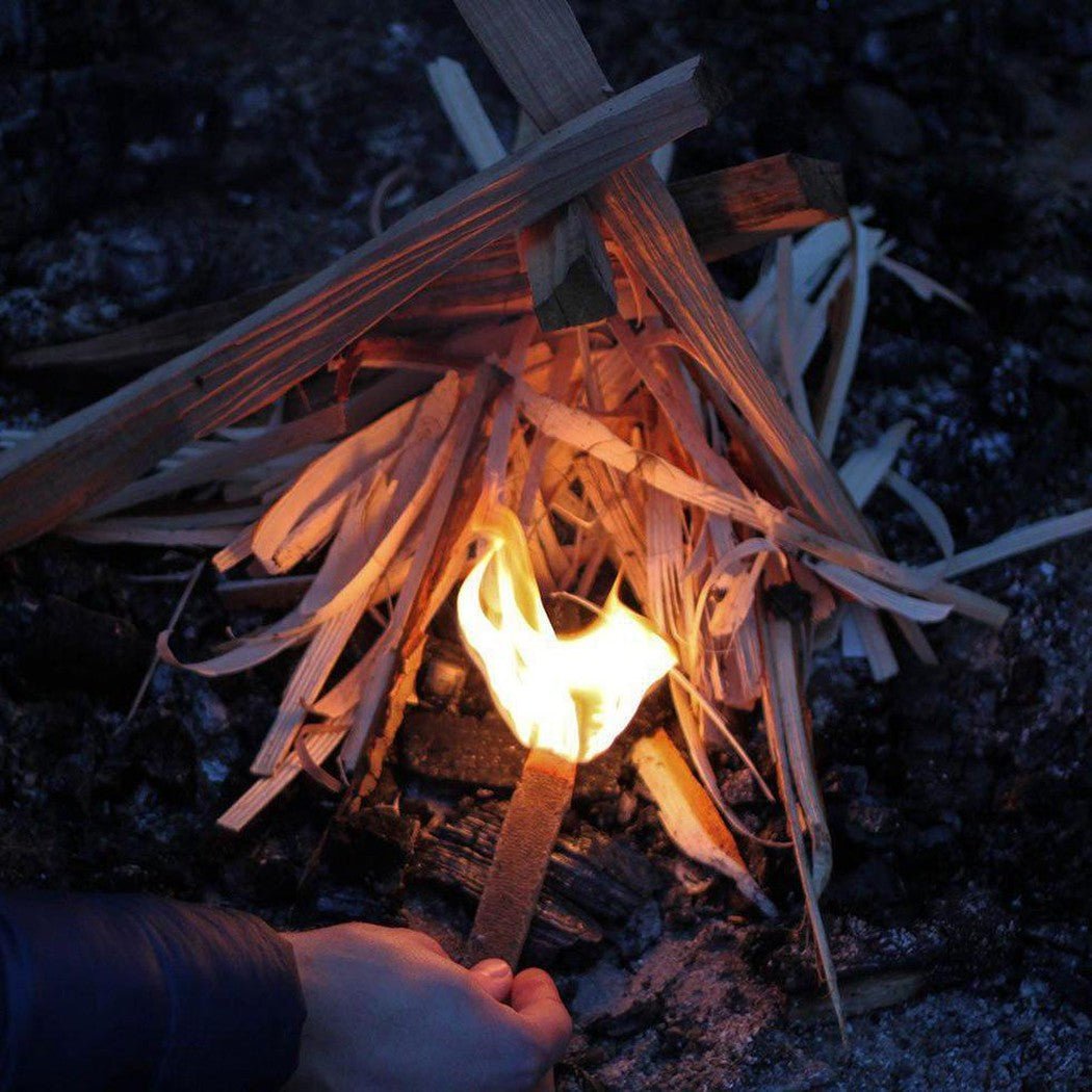 A hand holds the UCO Behemoth Sweetfire Match Kit near a small campfire made of wood strips in the dark.