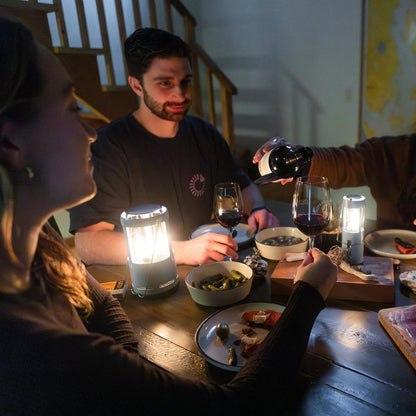 A woman and man sit at a table with two UCO Candlelier Candle Lanterns - Aluminum lit, as someone pours wine into a glass.