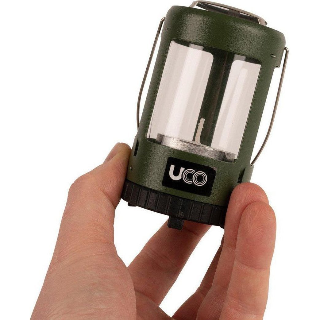 A hand holds the UCO Mini Lantern Kit 2.0, a green camping lantern with a clear front and black base, against a white background.