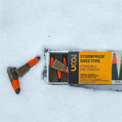 A box of UCO Stormproof Sweetfire fire starters sits in the snow with two pieces outside the box.