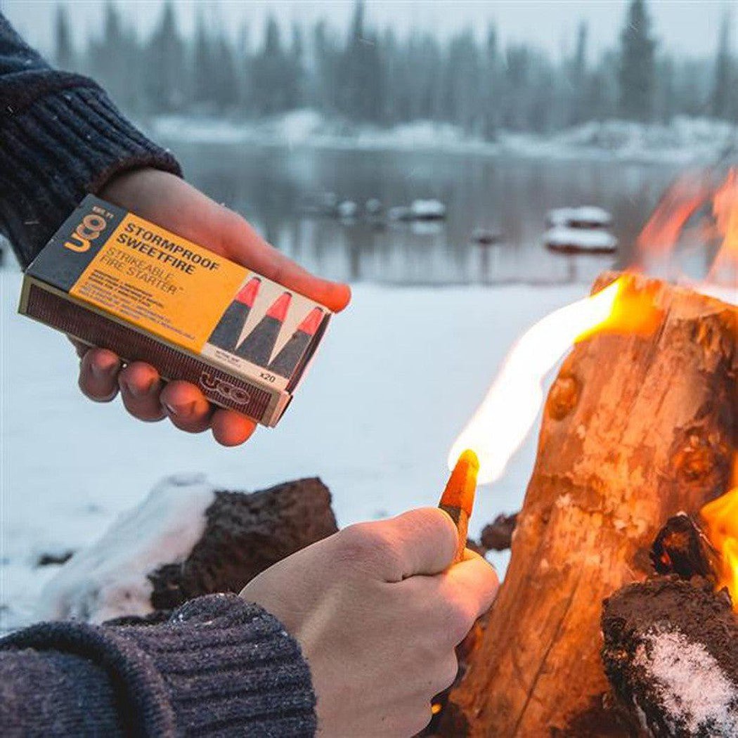 A hand holds a UCO Stormproof Sweetfire Firestarter box while lighting a match near a campfire by a snowy lake.