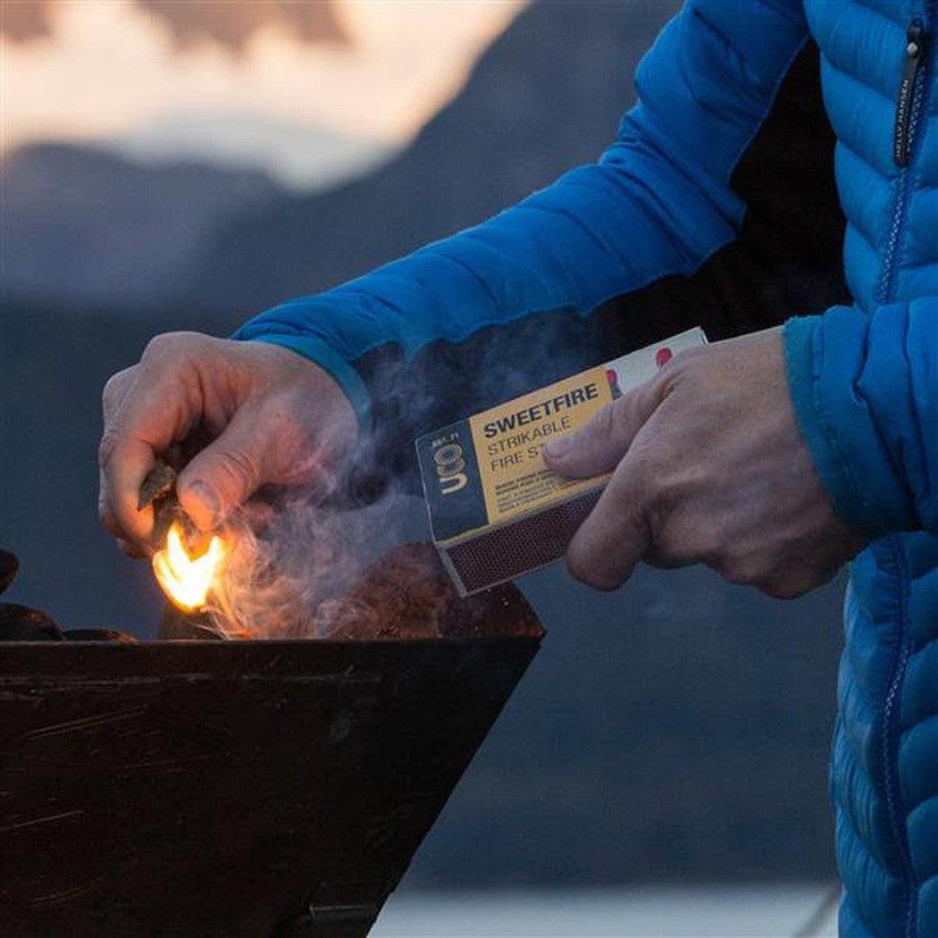 A person in a blue jacket lights a fire with UCO Sweetfire Strikeable Firestarter in a charcoal grill against a mountain backdrop.