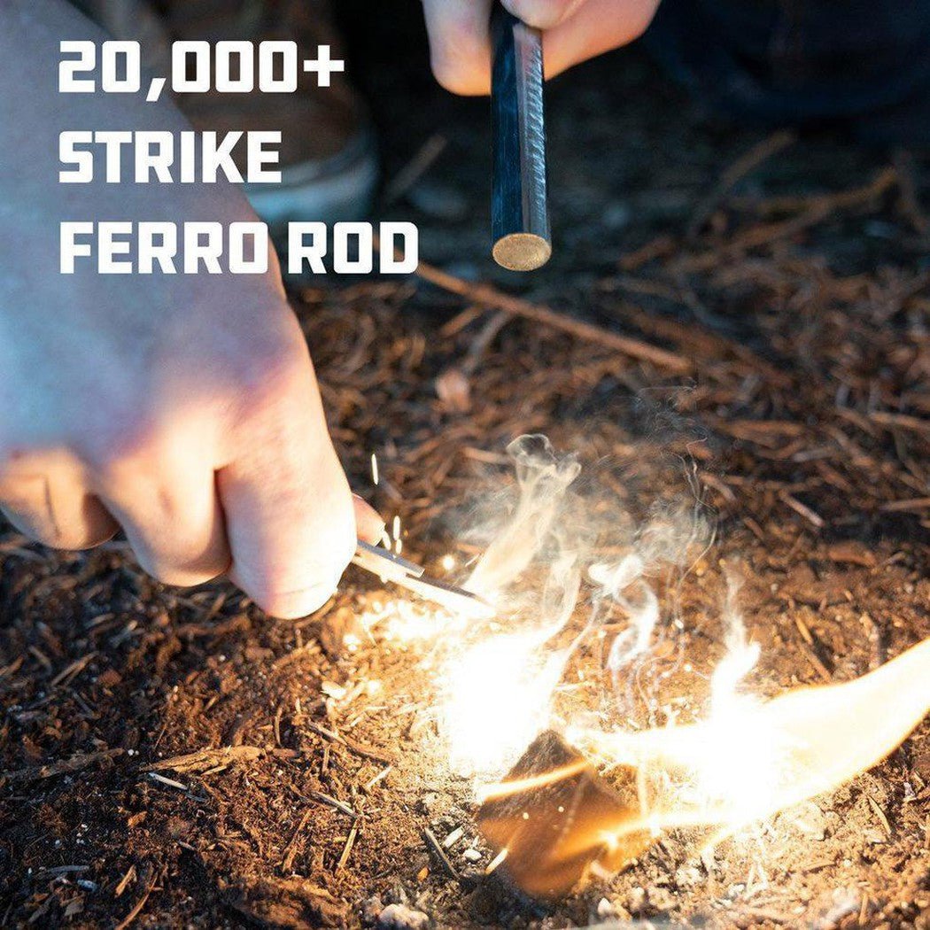 A hand strikes a ferro rod to start a fire on the ground with smoke and flames visible.